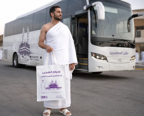 Pilgrim in white ihram standing beside a white bus, holding a white tote bag with purple Arabic text and a mosque illustration in front of him 
