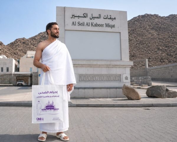 Man in white ihram robe stands outdoors, holding a white tote bag with purple Arabic text, beside a large stone sign reading Al Seil Al Kabeer Miqat under a blue sky 